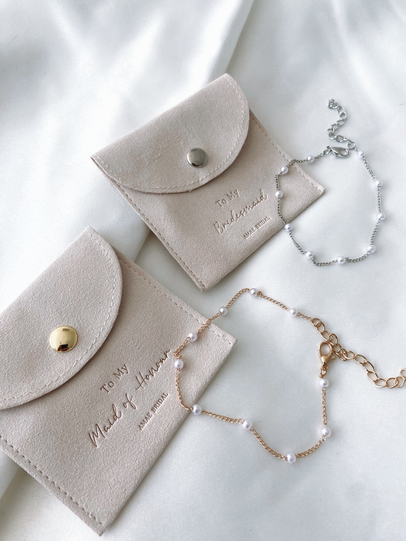 May include: Two light beige pouches with the text "To My Bridesmaid" and "To My Maid of Honor" embossed on them. Two pearl bracelets, one silver and one gold, are displayed next to the pouches. The bracelets have small, round pearls.