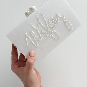 Peut inclure: Pochette rectangulaire blanche avec le nom "Wiley" &eacute;crit en perles. La pochette a un fermoir en perles et est tenue par une main portant des bracelets dor&eacute;s et argent&eacute;s.