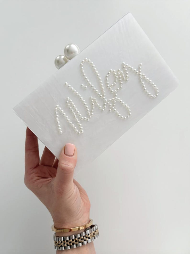 Peut inclure: Pochette rectangulaire blanche avec le nom "Wiley" &eacute;crit en perles. La pochette a un fermoir en perles et est tenue par une main portant des bracelets dor&eacute;s et argent&eacute;s.