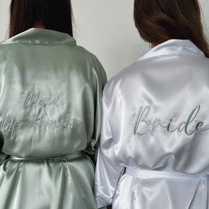 May include: Two satin robes, one sage green and one white, with embroidered text. The green robe says "Maid of Honor" and the white robe says "Bride".