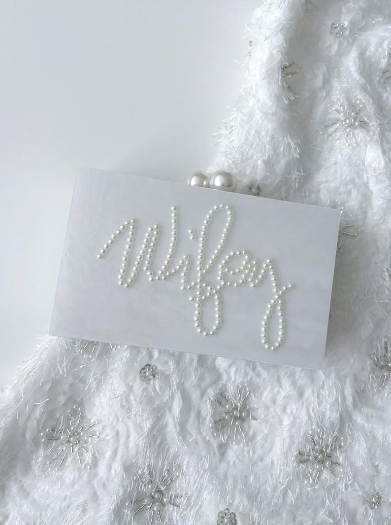 Peut inclure: Une pochette rectangulaire blanche avec le mot "Wifey" &eacute;pel&eacute; en perles blanches. La pochette a un fermoir en perle et repose sur un tissu blanc textur&eacute; avec des embellissements en forme de flocons de neige.