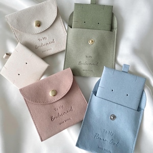 May include: Four small, square, velvet pouches in different colors: beige, green, pink, and blue. Each pouch has a gold button closure and a small opening at the top. The pouches are labeled with the text "To My Bridesmaid" or "To My Flower Girl" and the brand name "AMAR BRIDAL".