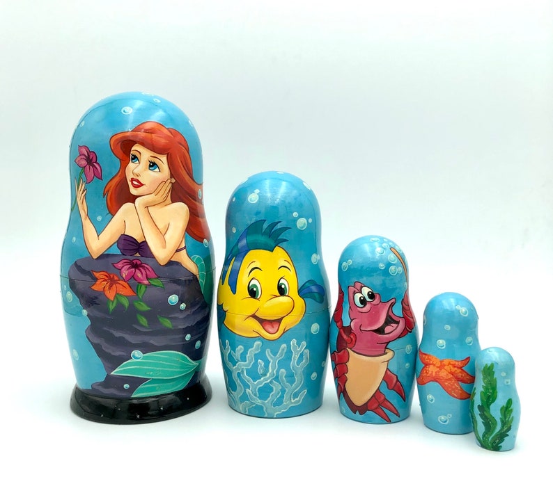 K&ouml;nnte beinhalten: Ein Satz von f&uuml;nf russischen Nistpuppen mit Figuren aus dem Disney-Film Die kleine Meerjungfrau. Die gr&ouml;&szlig;te Puppe zeigt Arielle, die Meerjungfrauenprinzessin, mit roten Haaren und einem gr&uuml;nen Schwanz. Die anderen Puppen zeigen Sebastian, Flounder und Ursula.