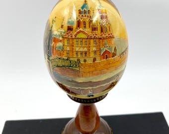 May include: A hand-painted wooden egg with a detailed depiction of a Russian church and surrounding buildings. The egg is displayed on a wooden stand with a gold-colored base.