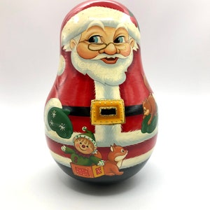 May include: A hand-painted wooden nesting doll of Santa Claus. The doll has a red coat, white beard, and a gold belt buckle. The base of the doll features illustrations of an elf, a fox, and a squirrel. A festive Christmas decoration.