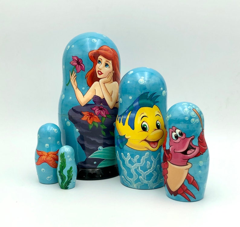 K&ouml;nnte beinhalten: Satz von f&uuml;nf Nistpuppen mit Figuren aus dem Disney-Film Die kleine Meerjungfrau. Die gr&ouml;&szlig;te Puppe zeigt Arielle, die Meerjungfrauenprinzessin, mit roten Haaren und einem gr&uuml;nen Schwanz. Die n&auml;chste Puppe zeigt Flounder, Arielles bester Freund, einen gelben Fisch mit blauen Flossen. Die dritte Puppe zeigt Sebastian, den Krebs, mit roten Krallen und einer violetten Schale. Die vierte Puppe ist ein rosa Krebs mit blauen Krallen. Die kleinste Puppe ist ein gelber Fisch mit blauen Flossen.