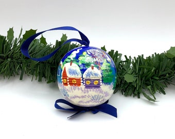May include: A blue and white Christmas ornament with a winter scene painted on it. The ornament features two small houses with snow-covered roofs and a snowy landscape.