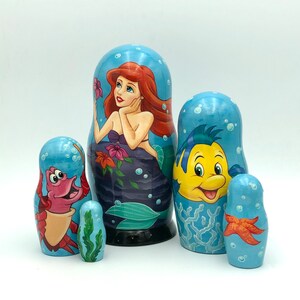 K&ouml;nnte beinhalten: Ein Set aus f&uuml;nf russischen Nistpuppen mit Figuren aus Disneys Arielle, die Meerjungfrau. Die gr&ouml;&szlig;te Puppe zeigt Arielle, die Meerjungfrauenprinzessin, mit langen roten Haaren und einem gr&uuml;nen Schwanz. Die n&auml;chste Puppe zeigt Flounder, Arielles besten Freund, einen gelben Fisch mit blauen Flossen. Die dritte Puppe ist Sebastian, der Krebs, mit roten Krallen und einer violetten Schale. Die vierte Puppe ist ein roter Krebs mit einer blauen Schale. Die kleinste Puppe ist ein Seestern mit einem gelben K&ouml;rper und orangefarbenen Flecken.