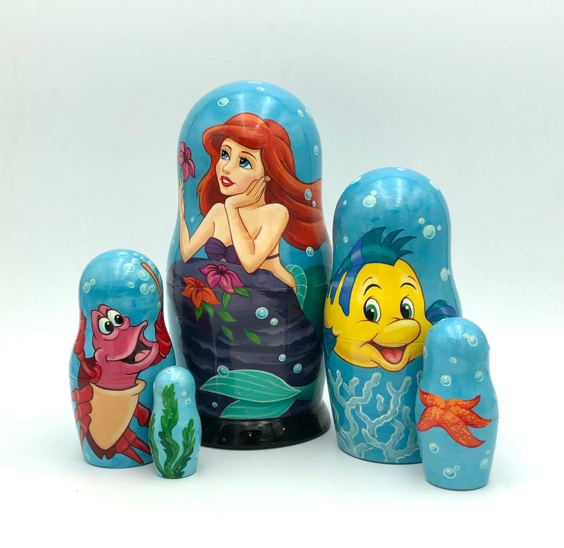K&ouml;nnte beinhalten: Ein Set aus f&uuml;nf russischen Nistpuppen mit Figuren aus Disneys Arielle, die Meerjungfrau. Die gr&ouml;&szlig;te Puppe zeigt Arielle, die Meerjungfrauenprinzessin, mit langen roten Haaren und einem gr&uuml;nen Schwanz. Die n&auml;chste Puppe zeigt Flounder, Arielles besten Freund, einen gelben Fisch mit blauen Flossen. Die dritte Puppe ist Sebastian, der Krebs, mit roten Krallen und einer violetten Schale. Die vierte Puppe ist ein roter Krebs mit einer blauen Schale. Die kleinste Puppe ist ein Seestern mit einem gelben K&ouml;rper und orangefarbenen Flecken.