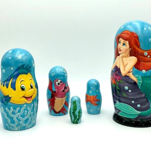 K&ouml;nnte beinhalten: Satz von f&uuml;nf Nistpuppen, oder Matroschka-Puppen, mit Figuren aus dem Disney-Film Die kleine Meerjungfrau. Die gr&ouml;&szlig;te Puppe zeigt Arielle, die Meerjungfrauenprinzessin, mit langen roten Haaren und einem lila Schwanz. Die anderen Puppen zeigen Flounder, Sebastian, einen Seestern und eine kleinere Arielle.