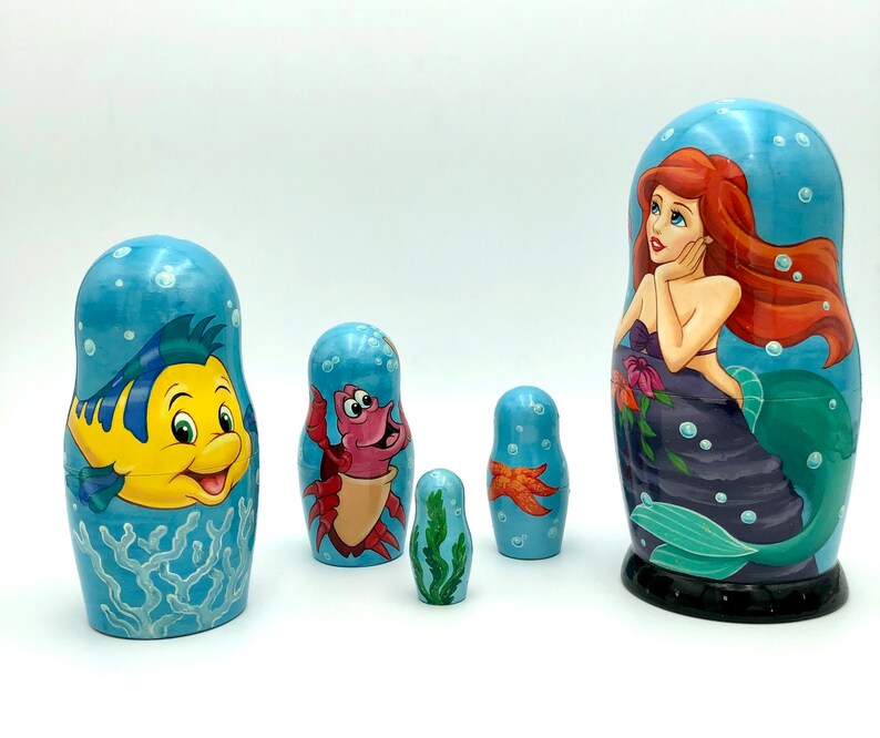 K&ouml;nnte beinhalten: Satz von f&uuml;nf Nistpuppen, oder Matroschka-Puppen, mit Figuren aus dem Disney-Film Die kleine Meerjungfrau. Die gr&ouml;&szlig;te Puppe zeigt Arielle, die Meerjungfrauenprinzessin, mit langen roten Haaren und einem lila Schwanz. Die anderen Puppen zeigen Flounder, Sebastian, einen Seestern und eine kleinere Arielle.