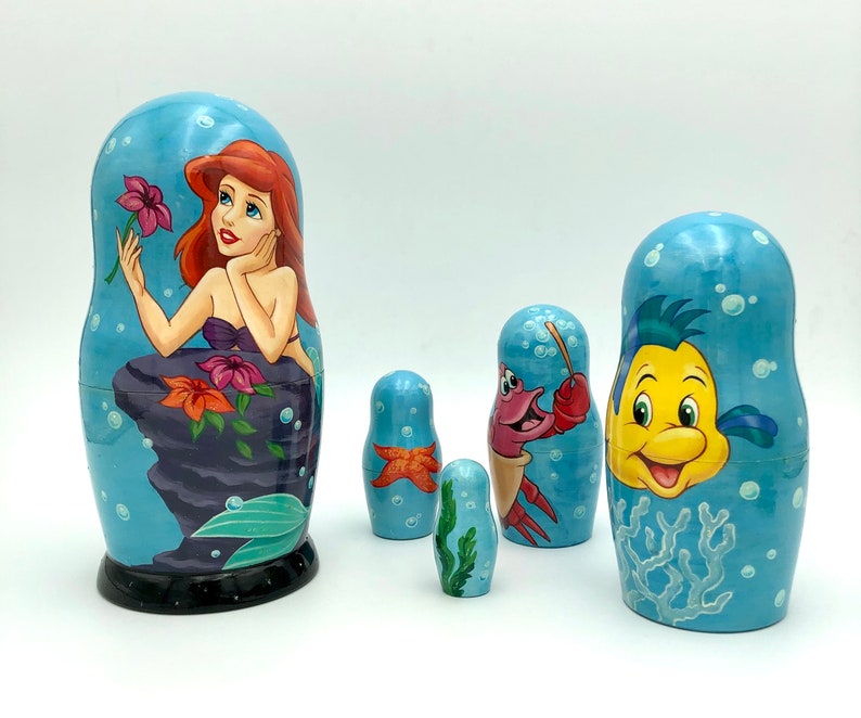 K&ouml;nnte beinhalten: Ein Satz von f&uuml;nf Nistpuppen, auch Matrjoschka-Puppen genannt, mit Figuren aus dem Disney-Film Die kleine Meerjungfrau. Die gr&ouml;&szlig;te Puppe zeigt Arielle, die Meerjungfrauenprinzessin, mit roten Haaren und einem lila Muschelbh.