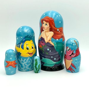 K&ouml;nnte beinhalten: Satz von f&uuml;nf Nistpuppen mit Figuren aus dem Disney-Film Die kleine Meerjungfrau. Die gr&ouml;&szlig;te Puppe zeigt Ariel, die Meerjungfrauenprinzessin, mit roten Haaren und einem lila Muschelbh. Die anderen Puppen zeigen Flounder, Sebastian und einen Seestern.
