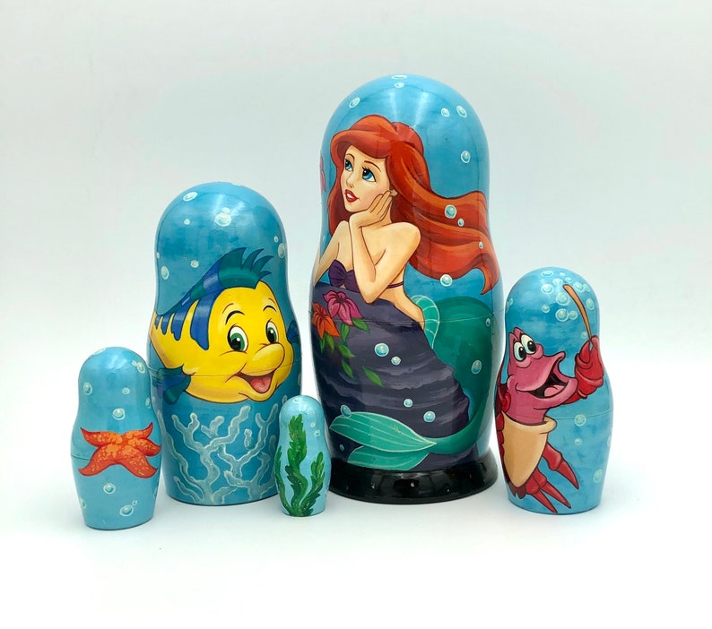 K&ouml;nnte beinhalten: Satz von f&uuml;nf Nistpuppen mit Figuren aus dem Disney-Film Die kleine Meerjungfrau. Die gr&ouml;&szlig;te Puppe zeigt Ariel, die Meerjungfrauenprinzessin, mit roten Haaren und einem lila Muschelbh. Die anderen Puppen zeigen Flounder, Sebastian und einen Seestern.