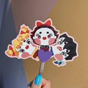 May include: Three cartoon character stickers with pink cheeks and large eyes. The characters are a girl with blonde hair, a girl with red hair and a boy with black hair. The stickers are attached to a purple heart-shaped charm.