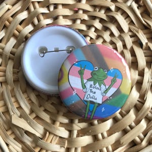 Puede incluir: Dos insignias redondas. Una muestra la parte posterior con un imperdible. La otra tiene un diseño colorido con un corazón y una rana que lleva una camiseta que dice "Protect The Dolls". El fondo es una cesta tejida.