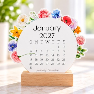 May include: A circular January 2027 calendar page featuring a colorful floral border with daisies, roses, poppies, and other blossoms. The calendar is mounted on a rectangular light wood base and includes the text January-Carnation.
