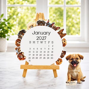 May include: A circular January 2027 calendar on a small wooden easel, surrounded by various dog breed illustrations. A small tan puppy figurine sits to the right of the easel, next to a rectangular base with the text ilamoo's WALL ART.