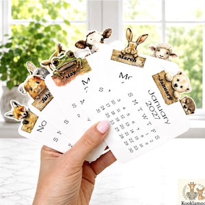 May include: A set of 2027 calendar cards featuring watercolor illustrations of animals including a frog, cow, rabbit, sheep, bear, and fox. Each card displays a month name on a wooden sign, with January 2027 clearly visible on the front card.