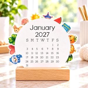 May include: A circular January 2027 calendar card set in a rectangular wooden base. The white card features black text and numbers, surrounded by colorful illustrations of a rabbit, flower, sun, star, watermelon, school bus, and snowman.