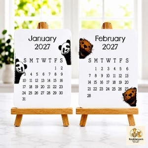 May include: Two white rectangular calendar cards for January and February 2027, each displayed on a small wooden easel. The January card features a panda illustration, while the February card displays an otter illustration, both with black text.