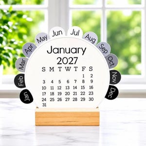 May include: A circular desk calendar featuring a white center with the text January 2027 and a grid of dates. Twelve semi-circular tabs labeled with the months of the year surround the perimeter. The calendar rests on a rectangular wooden base.