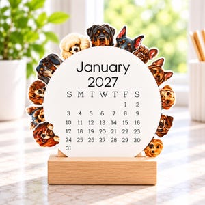 May include: A circular January 2027 calendar printed on white cardstock, featuring a border of various dog breeds. The calendar is held upright by a rectangular light-colored wooden base, displaying the month, year, and days of the month.