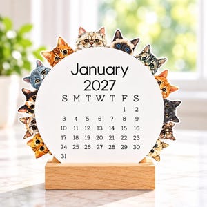 May include: A circular desk calendar for January 2027 featuring a white background with black text and numbers. The perimeter of the circle is decorated with various cat faces in different colors and patterns, all mounted on a rectangular wooden base.