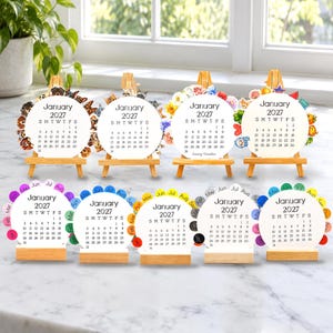 May include: Nine circular desk calendars for January 2027, each displayed on a small wooden easel or stand. The calendars feature black text on white backgrounds, with various colorful decorative borders including floral, animal, and seasonal motifs.