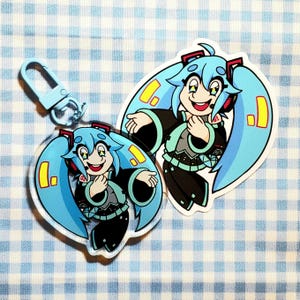 May include: Two blue and green anime character keychains with yellow exclamation points. The character has long blue hair and is wearing a black and green outfit. One keychain is attached to a silver keyring.