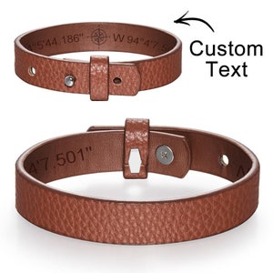 Puede incluir: Un brazalete de cuero marrón con cierre de hebilla. El brazalete tiene las coordenadas "4'5'44.186" W 94'4'7.5" grabadas en él. El brazalete también tiene el texto "Custom Text" grabado en él.
