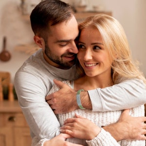 May include: A couple embracing, the man is wearing a light grey shirt and the woman is wearing a white sweater. The man is wearing a green and white bracelet on his wrist.