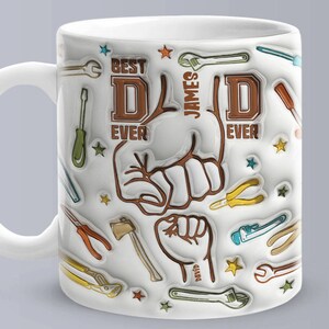 May include: White ceramic mug with a brown and white design. The mug features the text "Best Dad Ever" and "James" with a brown fist design. The mug is decorated with colorful tools and stars.