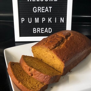 May include: A white letter board with the text "WELCOME GREAT PUMPKIN BREAD" and a loaf of pumpkin bread sliced on a white plate.