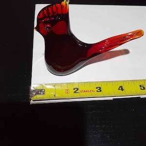 Può includere: Una statuetta di uccello in vetro stilizzata di colore rosso intenso con accenti arancioni e gialli sulla cresta e sulla coda. L'uccello è posizionato accanto a un metro a nastro giallo Stanley, che indica una lunghezza di circa 11,5 cm.