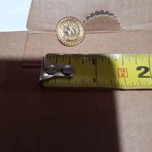 May include: A gold-colored button with a detailed design, approximately 1 inch in diameter, sits next to a yellow measuring tape. The button has a raised border and a central image. The tape measure shows the button's size.