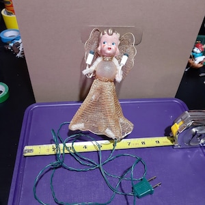 May include: A vintage angel tree topper with a gold mesh dress and a light-up face. The angel is holding two white lights in her hands. The angel is connected to a green wire with a plug.
