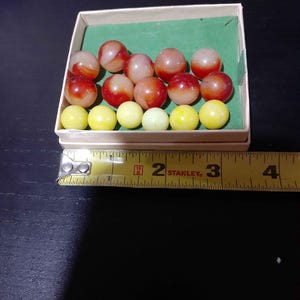 Puede incluir: Una colección de quince canicas de vidrio dispuestas en una caja de cartón rectangular con interior verde. El conjunto incluye nueve canicas rojas y blancas con remolinos y seis canicas amarillas sólidas. Una cinta métrica Stanley indica el ancho.