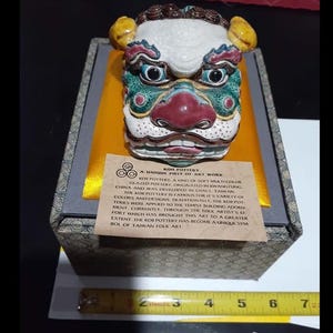 Pode incluir: Uma máscara de cerâmica pintada à mão, colorida, com uma expressão feroz, repousando sobre uma caixa forrada a cetim amarelo. A máscara apresenta detalhes em vermelho, verde, branco e amarelo. Uma pequena etiqueta de papel com texto é visível. A caixa tem cerca de 15 cm de comprimento.