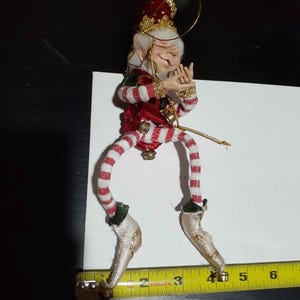May include: A decorative elf ornament with a red and green hat, red and white striped legs, and gold shoes. The elf has a smiling face and is wearing a red vest with gold trim. A yellow Stanley measuring tape shows the elf is 4.5 inches tall.