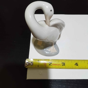 May include: A white porcelain swan figurine with a glossy finish, featuring a curved neck and a small black eye. The swan stands on a gray base with visible light pink feet. A yellow Stanley measuring tape shows the base is approximately 2 inches wide.