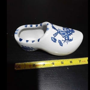 May include: A white ceramic Dutch clog with blue floral and windmill designs. The decorative shoe measures approximately 7 inches long. The open-topped shoe is a traditional Dutch design.