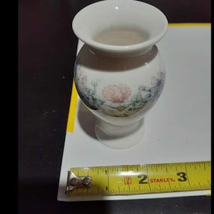May include: A small, white ceramic vase with a floral design. The vase is approximately 3 inches tall and has a rounded body with a flared top. The floral design features pink, blue, and green hues. The vase is on a white surface.