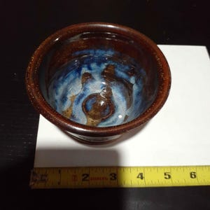 May include: A round ceramic bowl with a dark brown exterior and a glossy interior featuring a blend of blue, white, and brown glaze. A yellow measuring tape is positioned along the base, showing the bowl has a diameter of approximately 4 inches.