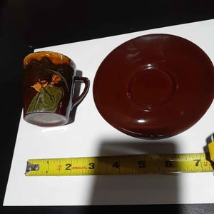 Puede incluir: Una taza y un plato de cerámica de color marrón oscuro. La taza presenta una figura pintada en relieve de una persona con capa y sombrero verdes tocando la flauta. Una cinta métrica amarilla Stanley muestra que el plato mide 17,78 cm de ancho.