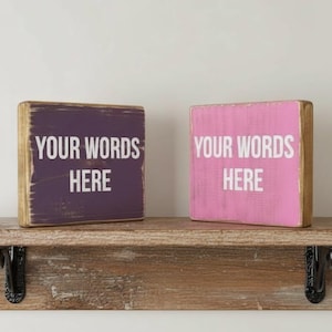 May include: Two square wooden blocks with distressed edges sit on a rustic wooden shelf with black metal brackets. The left block is dark purple and the right block is pink. Both feature the white text YOUR WORDS HERE in a sans-serif font.