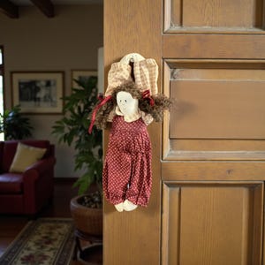 May include: A handmade doll door hanger. The doll has a white face, brown curly hair, and a red polka dot dress. The doll is attached to a cream-colored fabric with a floral pattern and red ribbons.