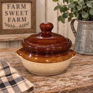 May include: A round ceramic bean pot with a glossy dark brown lid and upper rim, transitioning to a speckled cream-colored base. The pot features two small side handles and a central knob on the lid, showcasing a two-tone glazed finish.