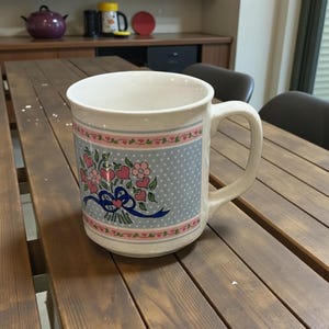 May include: White ceramic mug with a floral design featuring pink hearts and flowers, a blue ribbon, and a light blue polka dot background. The mug has a large handle and is sitting on a wooden table.