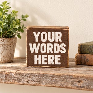 May include: A square wooden block sign with a dark brown stained finish and distressed edges. The front face features the white painted text "YOUR WORDS HERE" in a bold, sans-serif font, centered on the surface of the block.